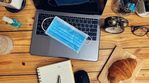 Schreibtisch unter anderem belegt von einem Laptop, einer Mouse, einem Wecker, einem Croissant und einer medizinischen Maske.