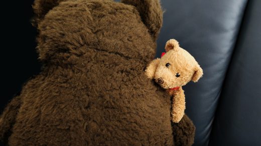 Ein kleiner beiger Teddybär liegt auf der rechten Schulter eines großen, braunen Teddybären.